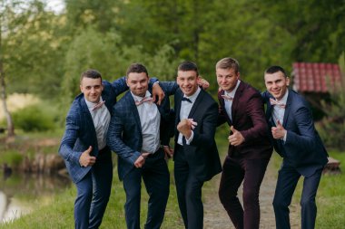 Takım elbiseli beş adamdan oluşan bir grup fotoğraf için poz veriyor ve bir kişi de onay veriyor. Sahne, erkekler giyinip fotoğraf çektirmek için poz verirken kutlama ve mutluluk dolu.