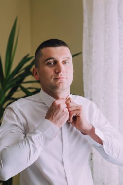 Beyaz gömlekli bir adam kravatını bağlamaya hazırlanıyor. Bir bitki ve pencerenin önünde duruyor.