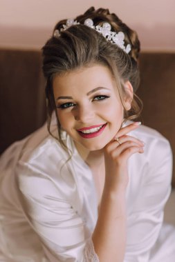 Beyaz cüppeli ve taclı bir kadın gülümsüyor. Kırmızı ruj sürmüş ve eli çenesinde.