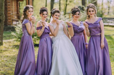 Mor elbiseli beş kadından oluşan bir grup beyaz elbiseli bir gelinle fotoğraf çektirmek için poz veriyor.