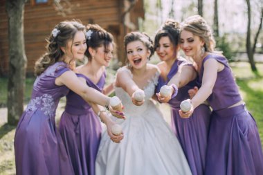 Bir grup kadın kapkeklerle fotoğraf çektirmek için poz veriyor. Gelin beyaz bir elbise giyiyor ve diğer kadınlar mor elbiseler giyiyor. Sahne mutlu ve kutlama dolu.