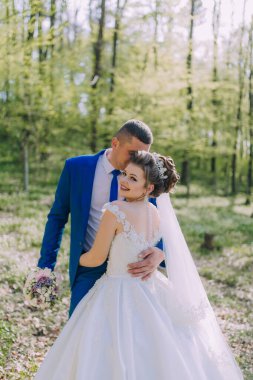 Bir gelin ve damat ormanda fotoğraf çektirmek için poz veriyorlar. Gelin beyaz bir elbise giyiyor ve damat mavi bir takım elbise giyiyor. İkisi de gülümsüyor ve birbirlerine sarılıyorlar.