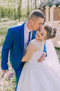 Bir gelin ve damat birbirlerinin yanaklarını öpüyor. Gelin beyaz bir elbise giyiyor ve damat mavi bir takım giyiyor.