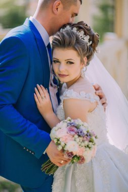 Mavi takım elbiseli ve mavi elbiseli bir gelin ve damat birbirlerine sarılıyorlar. Gelin elinde bir buket çiçek tutuyor.