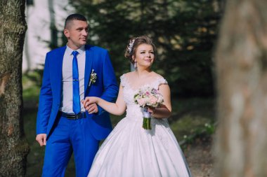 Bir gelin ve damat ormanda birlikte yürüyorlar. Gelin elinde bir buket çiçek tutuyor.