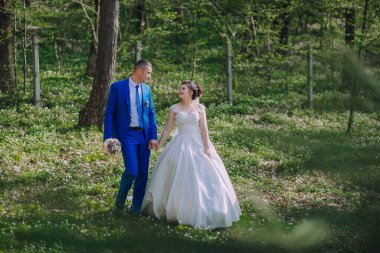 Bir gelin ve damat ormanda el ele tutuşup gülümsüyor. Gelin beyaz bir elbise giyiyor ve damat mavi bir takım elbise giyiyor. Sahne mutlu ve romantik.