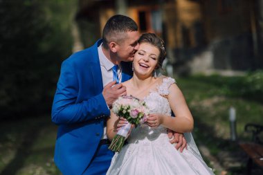 Bir gelin ve damat birbirlerine sarılıp öpüşüyorlar. Gelin elinde bir buket çiçek tutuyor.