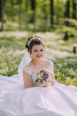 Beyaz elbiseli bir kadın elinde bir buket çiçekle bir tarlada oturuyor. Gülümsüyor ve mutlu.