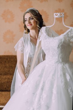 Bir gelin, askıda asılı beyaz bir elbisenin önünde duruyor. Gülümsüyor ve elbiseye bakıyor.