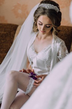 Kafasında beyaz bir peçeyle bir odada oturan bir gelin. Beyaz bir elbise ve beyaz çorap giyiyor.