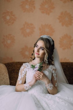 Bir gelin saçında çiçekle bir kanepede oturuyor. Beyaz bir elbise ve taç giyiyor.