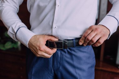 Bir adam beyaz gömlek ve mavi pantolon giyerken kemerini ayarlıyor. Formalite ve detaylara dikkat etme kavramı. Çünkü adam kıyafetini düzgün bir şekilde giydiğinden emin olmak istiyor.