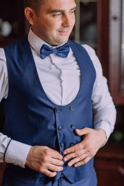 Mavi yelekli ve beyaz gömlekli bir adam papyon takıyor. Aynanın önünde duruyor, kravatını düzeltiyor.