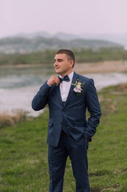 Mavi takım elbiseli ve beyaz gömlekli bir adam bir su kütlesinin yanında bir tarlada duruyor. Papyon ve yaka çiçeği takıyor.