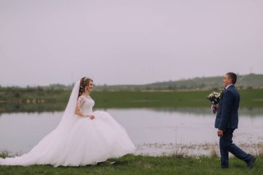 Gelin ve damat gölün kenarında duruyorlar, gelin beyaz bir elbise ve damat mavi bir takım elbise giyiyor. Gelin elinde bir buket çiçek tutuyor.