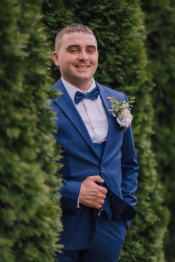 Mavi takım elbiseli ve beyaz gömlekli bir adam gülümsüyor ve elinde bir çiçek tutuyor. Bir çalılığın önünde duruyor, kolları çapraz ve başı eğik. Mutluluk ve memnuniyet kavramı