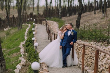 Bir gelin ve damat ormanda bir yolda duruyorlar. Gelin beyaz bir elbise giyiyor ve damat mavi bir takım elbise giyiyor. İkisi de gülümsüyor ve birbirlerine bakıyorlar.