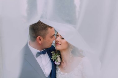 Bir gelin ve damat beyaz bir perdenin altında öpüşüyor. Gelin beyaz bir elbise giyiyor ve damat mavi bir takım giyiyor.