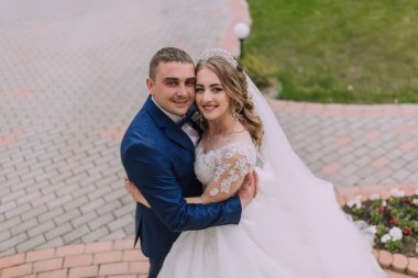 Bir gelin ve damat, tuğla kaldırımda fotoğraf çektirmek için poz veriyor. Gelin beyaz bir elbise giyiyor ve damat mavi bir takım elbise giyiyor. İkisi de gülümsüyor ve birlikte olmaktan mutlu görünüyorlar.