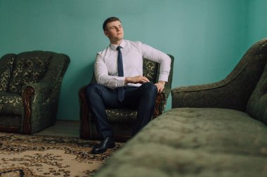 Takım elbiseli ve kravatlı bir adam diğer iki sandalyeyle aynı odada oturuyor. Birini ya da bir şeyi bekliyor.