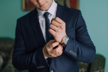 Takım elbiseli bir adam kravatını düzeltiyor ve saat takıyor. Formalite ve detaylara dikkat etme kavramı, adamın görünüşünün parlak ve profesyonel olması için zaman ayırması gibi.