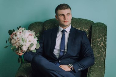 Takım elbiseli ve kravatlı bir adam elinde bir buket çiçekle yeşil bir sandalyede oturur. Zarafet ve entelektüellik anlayışı, çünkü adam resmi giyinmiş.