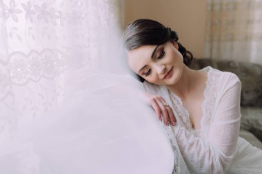 Beyaz elbiseli bir kadın kanepede oturuyor. Bir peçe takıyor ve başını çiçekli bir başlıkla örtüyor.