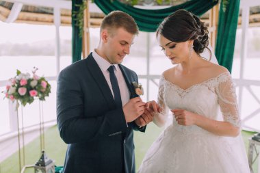 Bir erkek ve bir kadın evleniyor. Gelin beyaz bir elbise giyiyor ve damat mavi bir takım elbise giyiyor. Yeşil bir perdenin önünde duruyorlar.