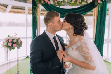 Yeşil bir kemerin önünde bir gelin ve damat öpüşüyor. Gelin beyaz bir elbise giyiyor ve damat mavi bir takım giyiyor.