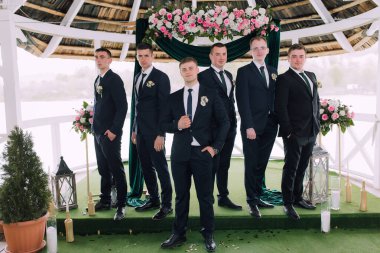 Takım elbiseli bir grup adam sahnede durup poz veriyor. Sahne, erkekler takım elbise ve kravat giydiği ve arka planda çiçekler olduğu için resmi ve kutlamalı.