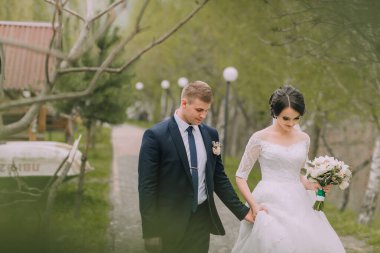 Bir gelin ve damat el ele tutuşup gülümsüyor. Gelin beyaz bir elbise giyiyor ve damat bir takım elbise giyiyor. Sahne ağaçlarla dolu bir parkta geçiyor.