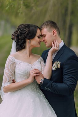 Bir gelin ve damat parkta birbirini kucaklıyor. Gelin beyaz bir elbise giyiyor ve damat mavi bir takım giyiyor.