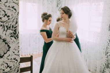 Bir gelin nedimelerinin yardımıyla düğününe hazırlanıyor. Gelin beyaz bir elbise ve duvak takıyor.