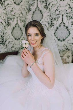 Bir gelin elinde bir çiçekle bir yatakta oturuyor. Beyaz bir elbise ve duvak takıyor.