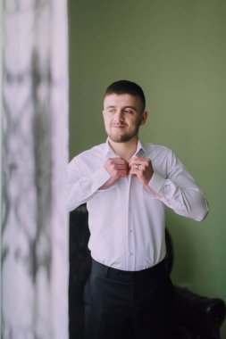 Beyaz gömlekli bir adam yeşil bir duvarın önünde giyiniyor. Siyah bir takım elbise giyiyor ve keyfi yerinde.