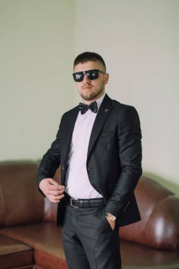 Takım elbiseli ve güneş gözlüklü bir adam kanepenin önünde duruyor. O bir kravat ve siyah bir ceket giyiyor.