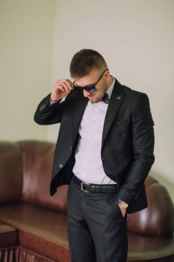 Takım elbiseli bir adam kanepenin önünde duruyor. Güneş gözlüğü ve kravat takıyor.