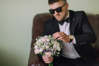 Takım elbiseli ve güneş gözlüklü bir adam elinde bir buket çiçek tutuyor. Deri bir koltukta oturuyor.