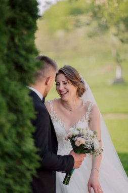 Bir gelin ve damat kameraya gülümsüyor. Gelin elinde bir buket çiçek tutuyor.