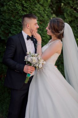 Bir gelin ve damat fotoğraf için poz veriyor. Gelin beyaz bir elbise giyiyor ve damat bir takım elbise giyiyor. Gelin elinde bir buket çiçek tutuyor.