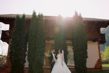 Önlerinde büyük bir ağaç olan bir binanın önünde bir gelin ve damat duruyor. Gelin beyaz bir elbise giyiyor ve damat siyah bir takım elbise giyiyor. Sahne mutlu ve kutlama dolu.