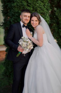 Bir gelin ve damat bir ağacın önünde fotoğraf için poz veriyorlar. Gelin beyaz bir elbise giyiyor ve damat bir takım elbise giyiyor. Elinde bir buket ve bir buket tutuyorlar.