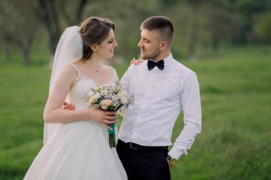 Gelin ve damat bir tarlada duruyorlar, gelin bir buket tutuyor ve damat siyah papyon takıyor.