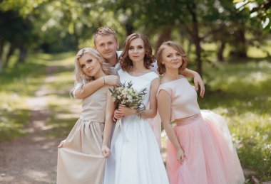 Bir gelin ve üç nedimesi ormanda fotoğraf çektirmek için poz veriyorlar. Gelin beyaz bir elbise giyiyor ve elinde bir buket tutuyor.