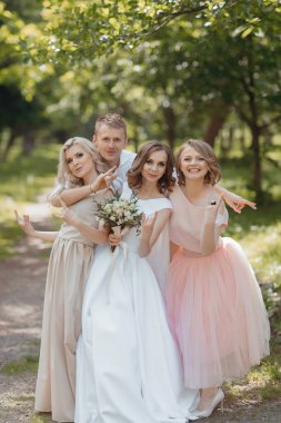 Bir gelin ve nedimeleri parkta fotoğraf çektirmek için poz veriyorlar. Gelin beyaz bir elbise giyip elinde buket tutarken diğer nedimeler pembe etek giyiyor.