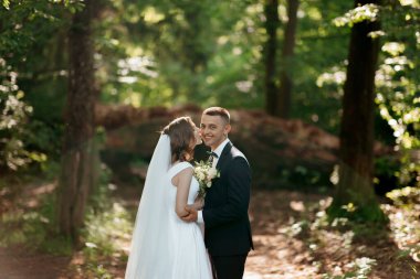 Gelin ve damat ormanda duruyorlar, gelin beyaz bir elbise giyiyor ve damat siyah bir takım elbise giyiyor. Birbirlerine gülümsüyor ve sarılıyorlar. Sıcak ve mutlu bir atmosfer yaratıyorlar.