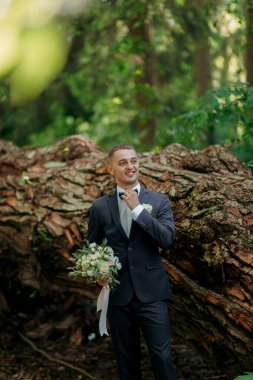 Takım elbiseli ve kravatlı bir adam büyük bir ağaç gövdesinin önünde duruyor. Elinde bir buket çiçek tutuyor ve gülümsüyor. Sahne romantik ya da samimi bir anı işaret ediyor.