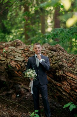 Takım elbiseli bir adam büyük bir ağaç gövdesinin yanında duruyor. Elinde bir buket çiçek ve beyaz bir fiyonk tutuyor. Sahne huzurlu ve huzurlu, mutlu ve memnun görünen bir adam.