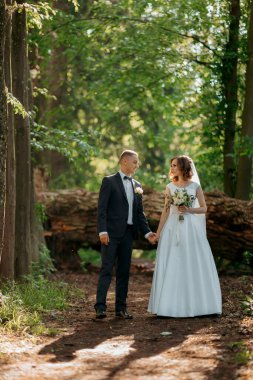 Bir gelin ve damat ormanda el ele tutuşup gülümsüyor. Gelin beyaz bir elbise giyiyor ve damat bir takım elbise giyiyor. Sahne huzurlu ve romantik.