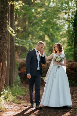 Bir gelin ve damat ormanda el ele tutuşup gülümsüyor. Gelin beyaz bir elbise giyiyor ve damat bir takım elbise giyiyor. Sahne huzurlu ve romantik.